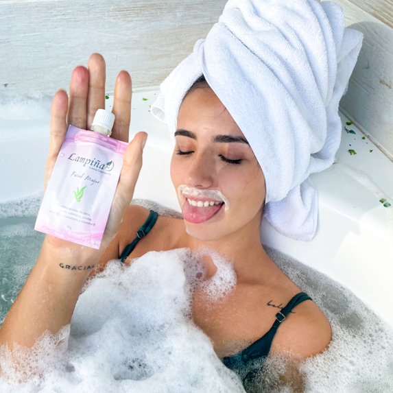 Mujer relajada en una bañera sosteniendo un sobre rosado de Crema Depilatoria Facial Lampiña de 15 g