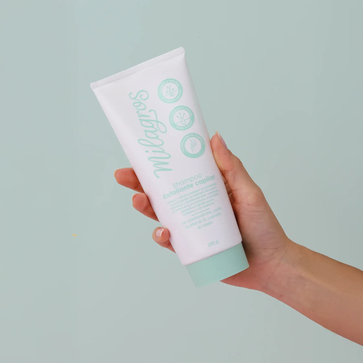Foto en mano → Mano sosteniendo tubo blanco Shampoo Exfoliante Capilar 200g Milagros con tapa menta, fondo verde agua
