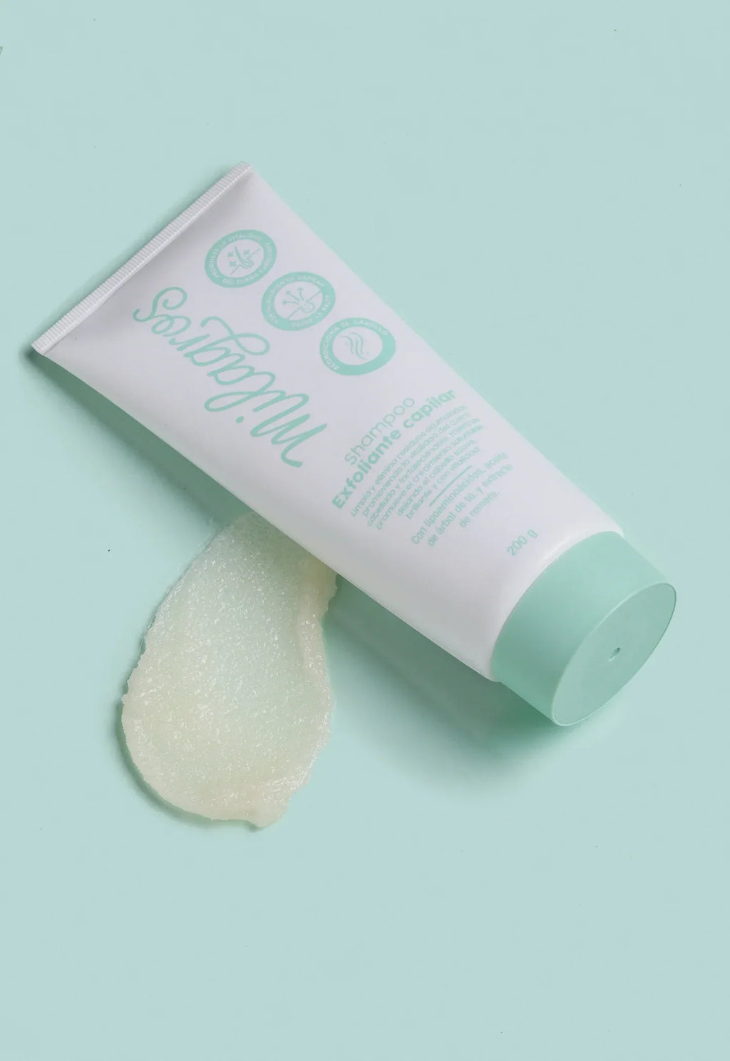 Tubo Shampoo Exfoliante Capilar Milagros 200g recostado sobre fondo verde agua junto a muestra de textura gel transparente con cristales de azúcar